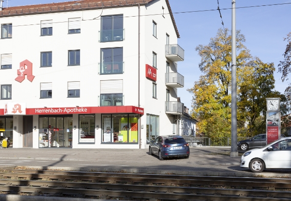 Foto des Gebäudes der Herrenbach-Apotheke mit nebenan liegendem Parkplatz für Kundinnen und Kunden
